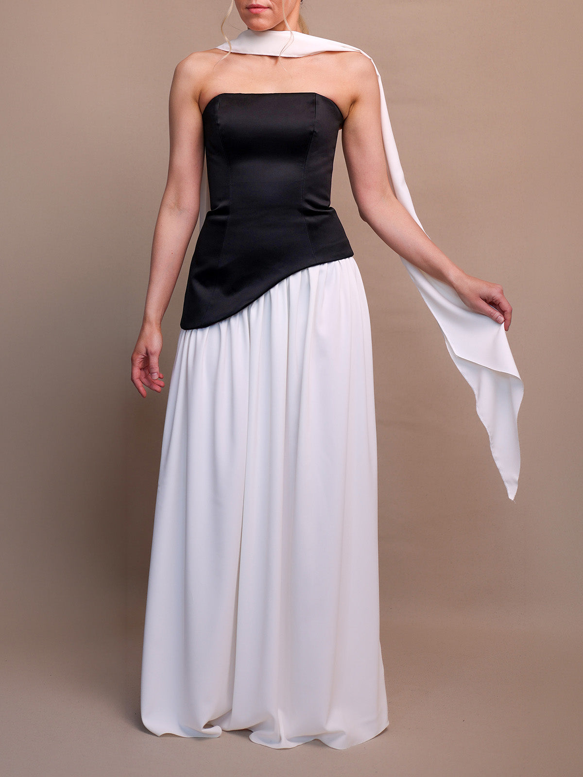 Woman wearing a black dress with asymmetric dropped waist and white skirt with a white scarf on a brown background. 